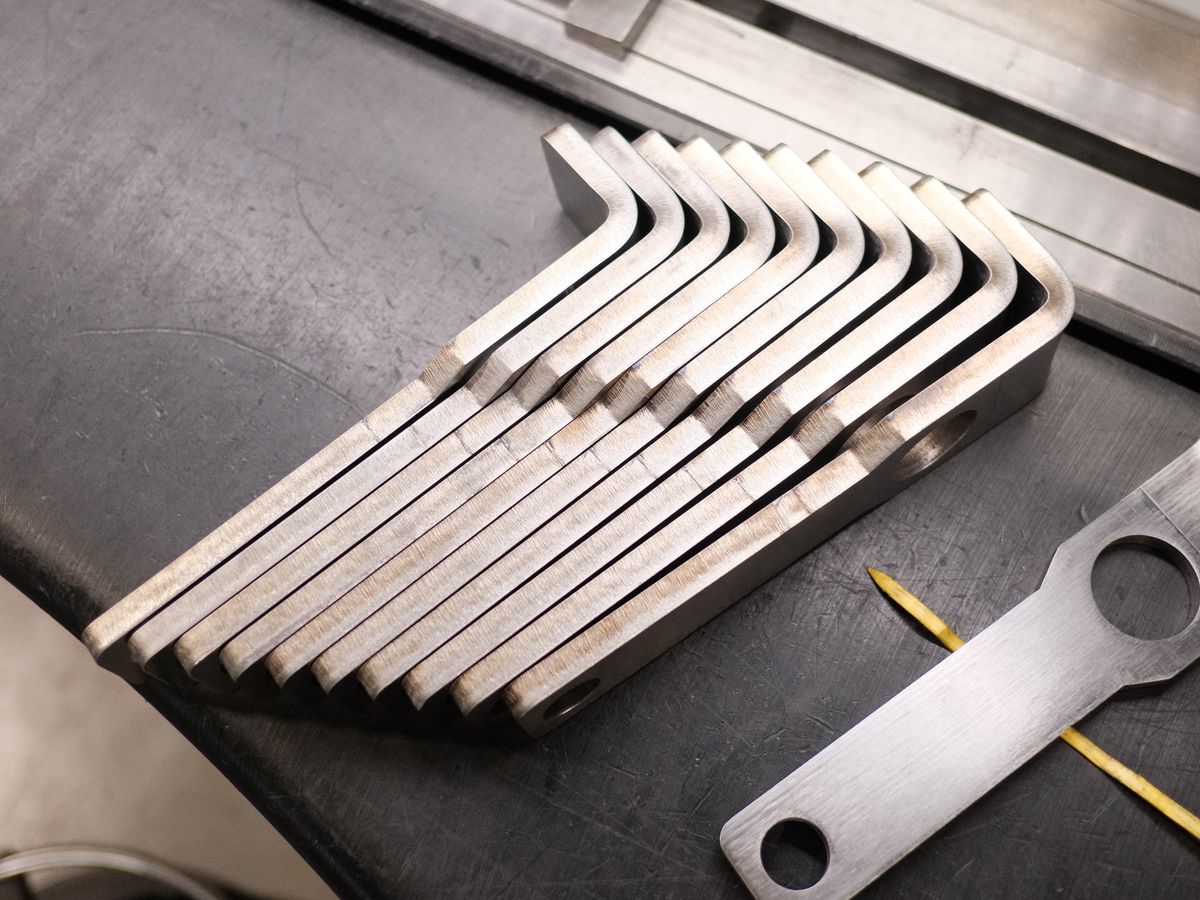 A fanned row of identical bent steel bars on the press brake bed beside a flat steel bracket at ACP Waterjet in Littleton, MA