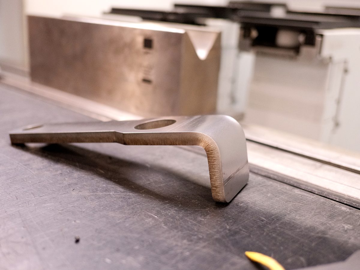 A bent bracket next to its flat blank on the press brake bed at ACP Waterjet