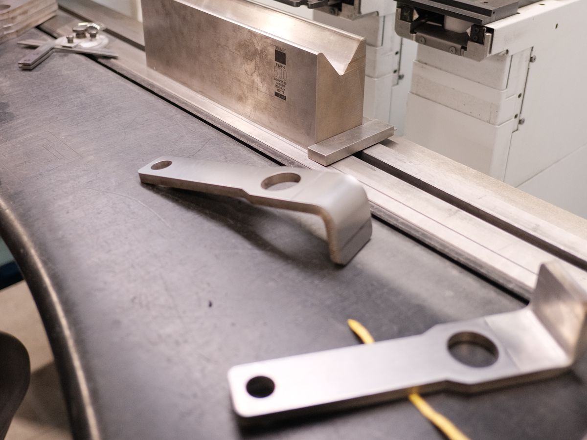 A bent steel bracket beside a flat cut bracket with holes, on the press brake bed at ACP Waterjet